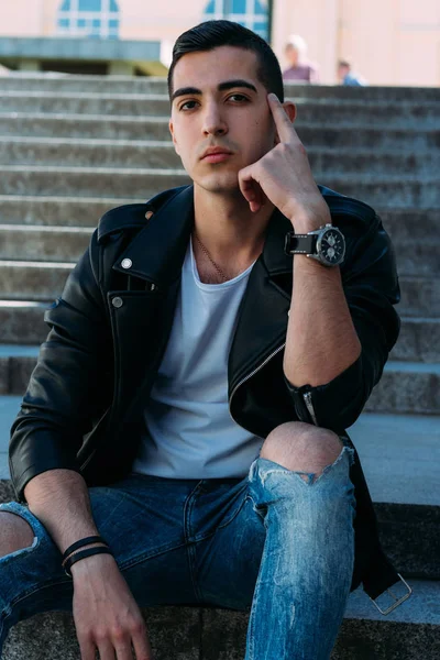 Seksi erkek poz korkuluk yakınındaki adımları oturur. çekici bir genç adam bir siyah deri ceket, beyaz bir gömlek ve beyaz spor ayakkabı. yandan izle