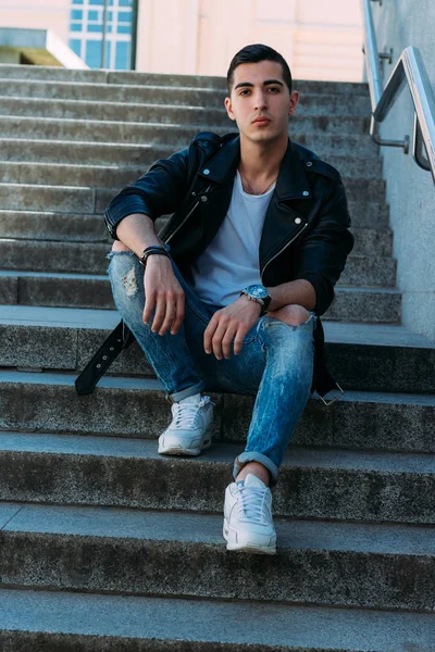 Seksi erkek poz korkuluk yakınındaki adımları oturur. çekici bir genç adam bir siyah deri ceket, beyaz bir gömlek ve beyaz spor ayakkabı. yandan izle