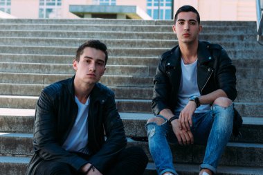 iki adam siyah ceket, beyaz T-shirt, kot pantolon ve Beyaz Ayakkabı içinde belgili tanımlık merdiven üstünde oturuyorsun. birde ellerinde. sosyalleşme, Toplantı arkadaşlar. parmaklıklar yakınındaki şehirde