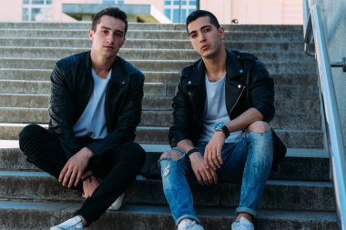 iki adam siyah ceket, beyaz T-shirt, kot pantolon ve Beyaz Ayakkabı içinde belgili tanımlık merdiven üstünde oturuyorsun. birde ellerinde. sosyalleşme, Toplantı arkadaşlar. parmaklıklar yakınındaki şehirde