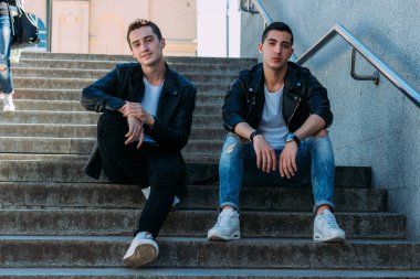 iki adam siyah ceket, beyaz T-shirt, kot pantolon ve Beyaz Ayakkabı içinde belgili tanımlık merdiven üstünde oturuyorsun. birde ellerinde. sosyalleşme, Toplantı arkadaşlar. parmaklıklar yakınındaki şehirde