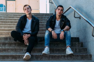 iki adam siyah ceket, beyaz T-shirt, kot pantolon ve Beyaz Ayakkabı içinde belgili tanımlık merdiven üstünde oturuyorsun. birde ellerinde. sosyalleşme, Toplantı arkadaşlar.