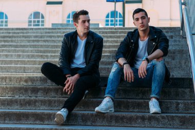 iki adam siyah ceket, beyaz T-shirt, kot pantolon ve Beyaz Ayakkabı içinde belgili tanımlık merdiven üstünde oturuyorsun. birde ellerinde. sosyalleşme, Toplantı arkadaşlar.