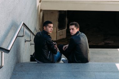 iki adam siyah ceket, kot pantolon ve Beyaz Ayakkabı içinde belgili tanımlık merdiven üstünde oturuyorsun. birde ellerinde. sosyalleşme, Toplantı arkadaşlar.