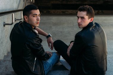 iki adam siyah ceket, kot pantolon ve Beyaz Ayakkabı içinde belgili tanımlık merdiven üstünde oturuyorsun. birde ellerinde. sosyalleşme, Toplantı arkadaşlar.