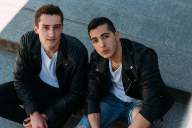 iki adam siyah ceket, kot pantolon ve Beyaz Ayakkabı içinde belgili tanımlık merdiven üstünde oturuyorsun. birde ellerinde. sosyalleşme, Toplantı arkadaşlar.