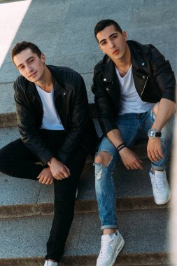 iki adam siyah ceket, kot pantolon ve Beyaz Ayakkabı içinde belgili tanımlık merdiven üstünde oturuyorsun. birde ellerinde. sosyalleşme, Toplantı arkadaşlar.