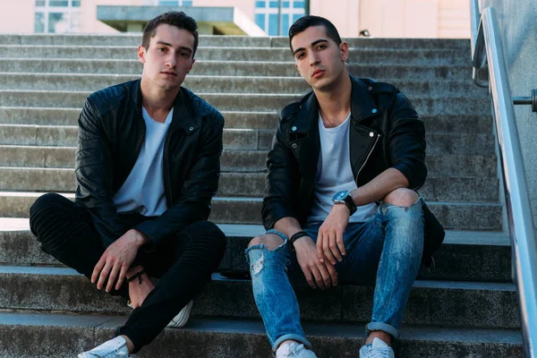 iki adam siyah ceket, beyaz T-shirt, kot pantolon ve Beyaz Ayakkabı içinde belgili tanımlık merdiven üstünde oturuyorsun. birde ellerinde. sosyalleşme, Toplantı arkadaşlar. parmaklıklar yakınındaki şehirde