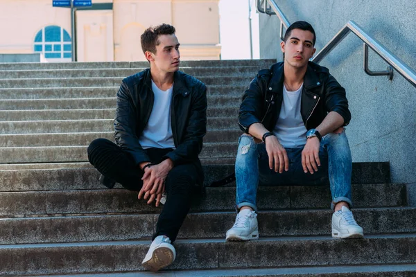 Dos tipos con chaquetas negras, camisetas blancas, jeans y zapatos blancos están sentados en las escaleras. en las manos de un reloj. socializar, conocer amigos . — Foto de Stock
