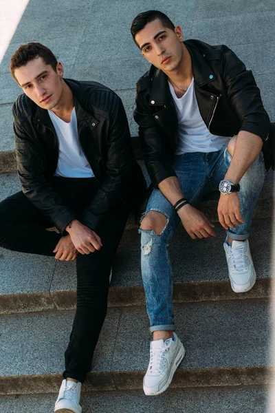 Dos tipos en chaquetas negras, jeans y zapatos blancos están sentados en las escaleras. en las manos de un reloj. socializar, conocer amigos . — Foto de Stock