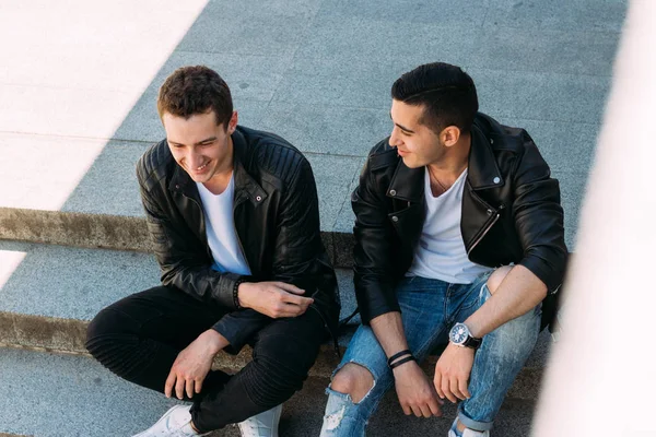 Deux gars en vestes noires, jeans et chaussures blanches, en T-shirts blancs sont assis sur les marches. sur les mains d'une montre. socialiser, rencontrer des amis . — Photo