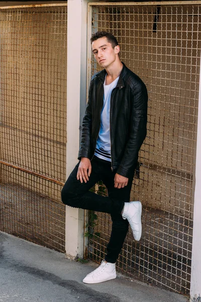 güzel ve çekici adam şehir kılavuzunda yakın duran, poz. siyah deri kurta, beyaz T-shirt, kot pantolon ve beyaz ayakkabı giymiş.