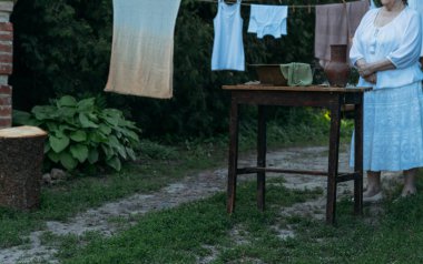 Yaşlı bir kadın duruyor ve tablo yakınındaki avluda beyaz vintage giysiler içinde bekler. Eski bir kil sürahi ve eski bir bakır lavabo su ve bez ile tablodur. sonraki clothespins vardır. Asılı, giysi kurutulur. köy yıkama.