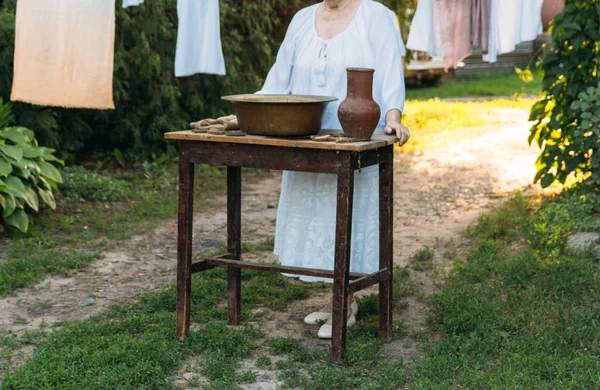 Yaşlı bir kadın duruyor ve tablo yakınındaki avluda beyaz vintage giysiler içinde bekler. Eski bir kil sürahi ve eski bir bakır lavabo su ve bez ile tablodur. sonraki clothespins vardır. Asılı, giysi kurutulur. köy yıkama.