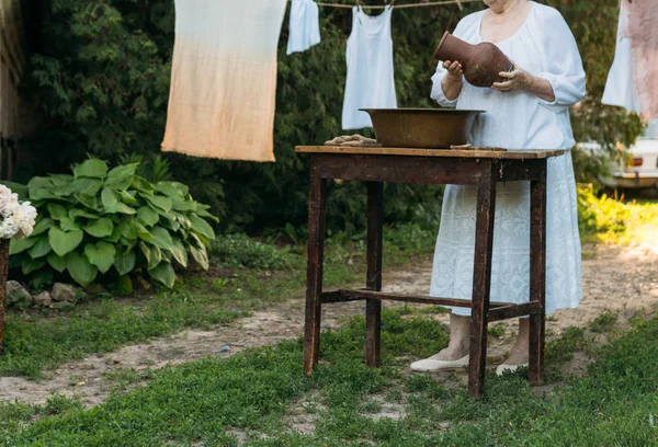 Yaşlı bir kadın bahçede giysi yıkama. Beyaz vintage giyim. Taşra hayatı. elbise bir ip üzerinde kurur. Masanın yanındaki clothespins vardır. Eski bir bakır lavabo ve kil sürahi Stark. kadın su dökülen