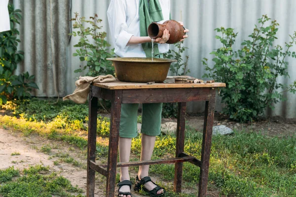 beyaz bir gömlek ve yeşil kazak, çocukta bir kil sürahi tutar, suyun dışında bakır lavabo içine dökülen. Yardım, kırsal. Köyü.