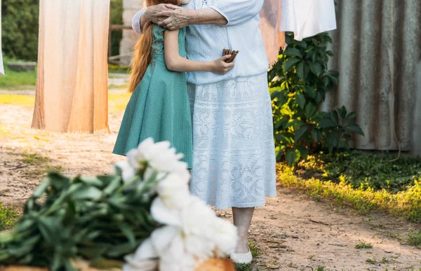 a grandmother, an elderly woman in white, vintage clothes stands and hugs her granddaughter, a girl in a green dress, in the garden. meeting and congratulations, a gift. on the stump is a bouquet of white peonies. in the background,