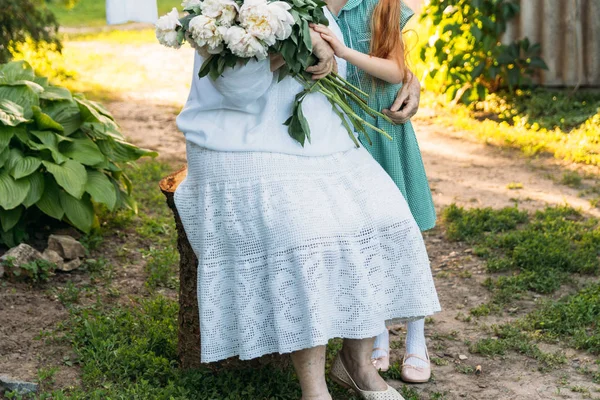 ヴィンテージ服を着て座っている彼女の孫娘 緑のドレスの少女を抱き締めると 庭の花 白牡丹の花束他の手を握って彼女の年配の女性 会議やお祝い 贈り物 — ストック写真