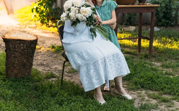 grandmother, an elderly woman in white, vintage clothes sitting and hugging her granddaughter, a girl in a green dress, and holding in her other hand a bouquet of flowers, white peonies, in the garden. meeting and congratulations, a gift
