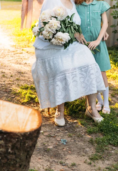 ヴィンテージ服を着て座っている彼女の孫娘 緑のドレスの少女を抱き締めると 庭の花 白牡丹の花束他の手を握って彼女の年配の女性 会議やお祝い 贈り物 — ストック写真