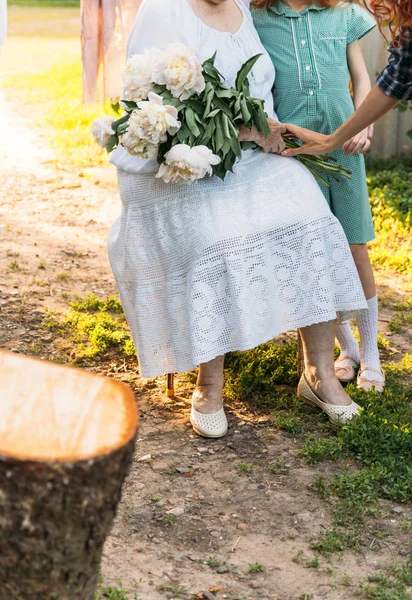 grandmother, an elderly woman in white, vintage clothes sitting and hugging her granddaughter, a girl in a green dress, and holding in her other hand a bouquet of flowers, white peonies, in the garden. meeting and congratulations, a gift.a female han