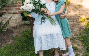 Aile bağları. Büyükanne, yaşlı bir kadın oturuyor ve onun torunu, Yeşil elbiseli bir kız sarılma ve onu bir buket çiçek, beyaz peonies, bahçedeki diğer elini tutarak beyaz, vintage giysiler içinde. Toplantı ve tebrik ederim, bir hediye