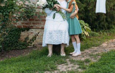 Büyüklerin saygı. Beyaz vintage giysiler içinde yaşlı bir kadın bahçede bir sandalyeye oturur ve bir buket beyaz peonies onun elinde tutar. yeşil giysili, onun büyükannesi sarılarak torunu yan.