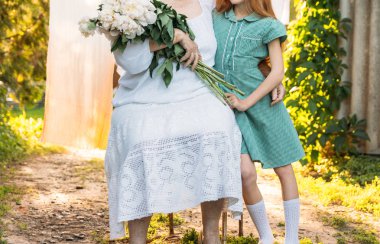 Büyüklerin saygı. Toplantı akrabaları. Büyükanne, yaşlı bir kadın oturuyor ve onun torunu, Yeşil elbiseli bir kız sarılma ve onu bir buket çiçek, beyaz peonies, bahçedeki diğer elini tutarak beyaz, vintage giysiler içinde. Toplantı