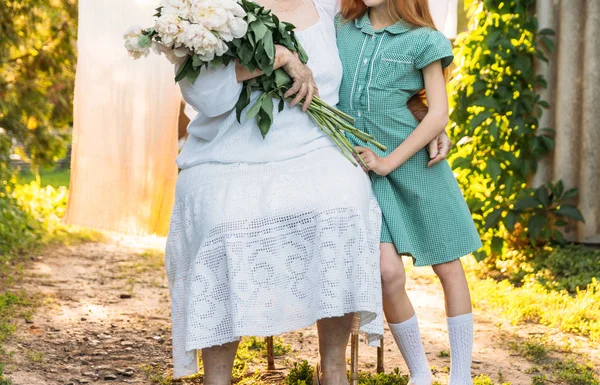 Büyüklerin saygı. Toplantı akrabaları. Büyükanne, yaşlı bir kadın oturuyor ve onun torunu, Yeşil elbiseli bir kız sarılma ve onu bir buket çiçek, beyaz peonies, bahçedeki diğer elini tutarak beyaz, vintage giysiler içinde. Toplantı