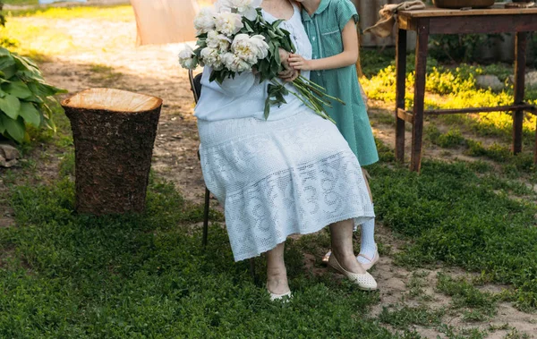 grandmother, an elderly woman in white, vintage clothes sitting and hugging her granddaughter, a girl in a green dress, and holding in her other hand a bouquet of flowers, white peonies, in the garden. meeting and congratulations, a gift