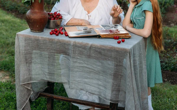 Büyükanne torunu ile masada oturur, onun gençlik fotoğrafları ile eski bir albüm onu gösteriyor. bir kil vazo çiçek ve kiraz ile geçerli. Aile öyküleri ve anılar. Yaşlı kadın vintage gelinlik ve kız giymiş ben