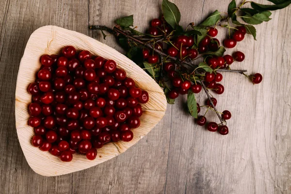 fresh red cherries in a wooden plate on a wooden table. wooden plate on a wooden background. next to it is a cherry branch with cherries and green leaves.a plate in the form of a triangle. ecology. proper nutrition.