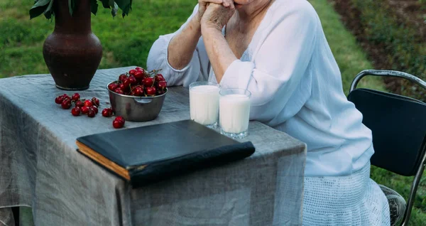 Emlékek az idősek. a nagymamája ül egy széken, a kertben. az asztalon van, két pohár tej, egy tányér, és a cseresznye virágok egy agyag váza, egy régi albumot, vagy egy könyv — Stock Fotó