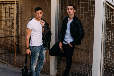 güzel ve çekici adam şehirde ayakta, poz. siyah deri kurta, beyaz T-shirt, kot pantolon ve beyaz ayakkabı giymiş.