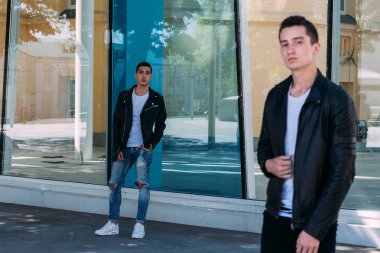 güzel ve çekici adam şehirde ayakta, poz. siyah deri kurta, beyaz T-shirt, kot pantolon ve beyaz ayakkabı giymiş. ayna Binası yakınındaki