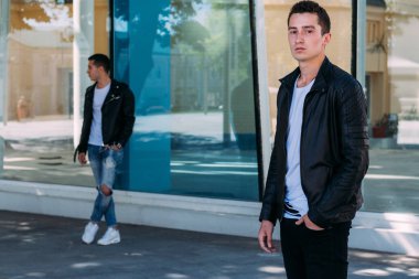 güzel ve çekici adam şehirde ayakta, poz. siyah deri kurta, beyaz T-shirt, kot pantolon ve beyaz ayakkabı giymiş. ayna Binası yakınındaki