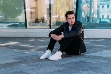 güzel ve çekici oturur yerde şehirde bina, poz bir cam yakınındaki biri. siyah deri kurta, beyaz T-shirt, kot pantolon ve beyaz ayakkabı giymiş. Şehir stili