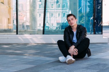 güzel ve çekici oturur yerde şehirde bina, poz bir cam yakınındaki biri. siyah deri kurta, beyaz T-shirt, kot pantolon ve beyaz ayakkabı giymiş. Şehir stili