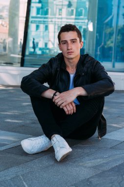 güzel ve çekici oturur yerde şehirde bina, poz bir cam yakınındaki biri. siyah deri kurta, beyaz T-shirt, kot pantolon ve beyaz ayakkabı giymiş. Şehir stili
