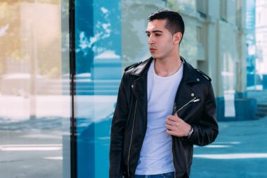 güzel ve çekici adam şehirde bina, poz cam duruyor. siyah deri kurta, beyaz T-shirt, kot pantolon ve beyaz ayakkabı giymiş.