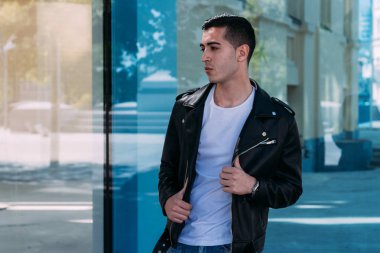 güzel ve çekici adam şehirde bina, poz cam duruyor. siyah deri kurta, beyaz T-shirt, kot pantolon ve beyaz ayakkabı giymiş.