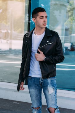 güzel ve çekici adam şehirde bina, poz cam duruyor. siyah deri kurta, beyaz T-shirt, kot pantolon ve beyaz ayakkabı giymiş.