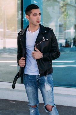 güzel ve çekici adam şehirde bina, poz cam duruyor. siyah deri kurta, beyaz T-shirt, kot pantolon ve beyaz ayakkabı giymiş.