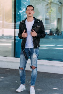 güzel ve çekici adam şehirde bina, poz cam duruyor. siyah deri kurta, beyaz T-shirt, kot pantolon ve beyaz ayakkabı giymiş.