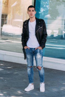 güzel ve çekici adam şehirde bina, poz cam duruyor. siyah deri kurta, beyaz T-shirt, kot pantolon ve beyaz ayakkabı giymiş.