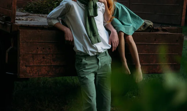 a barefooted girl in a green, vintage dress, the dress sits on the back of a red, old car and hugs the barefoot neck, next to a standing guy who is dressed in a white shirt, green shorts with a green sweater tied around his neck. summer day in the vi