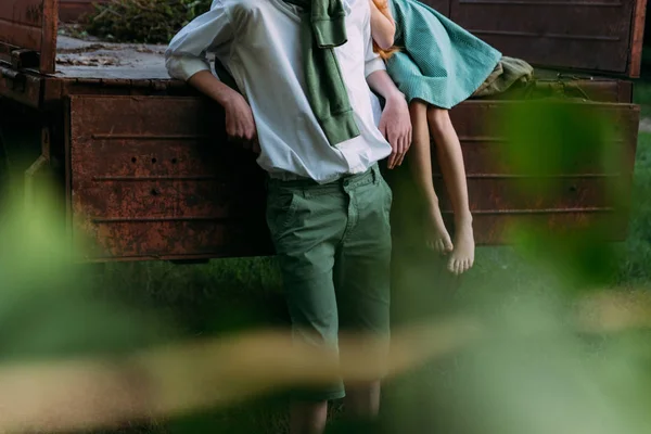 to look from behind bushes. blurred leaves at the beginning of the snapshot. a barefooted girl in a green, vintage dress, the dress sits on the back of a red, old car and hugs the barefoot neck, next to a standing guy who is dressed in a white shirt,