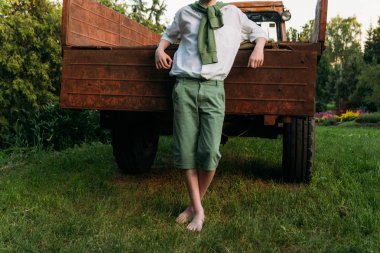 a boy in a white shirt and green pants, a green sweater around his neck, leaning his elbows on the old truck cousin, barefoot on the grass in the summer. village, farmer, on farm. hands in pockets clipart