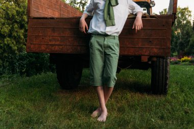 yeşil çimenlerin üzerinde durmak için çıplak ayak. adam beyaz bir gömlek ve bir yeşil kazak eski bir kırmızı traktör gövdesi yakınındaki yeşil şort