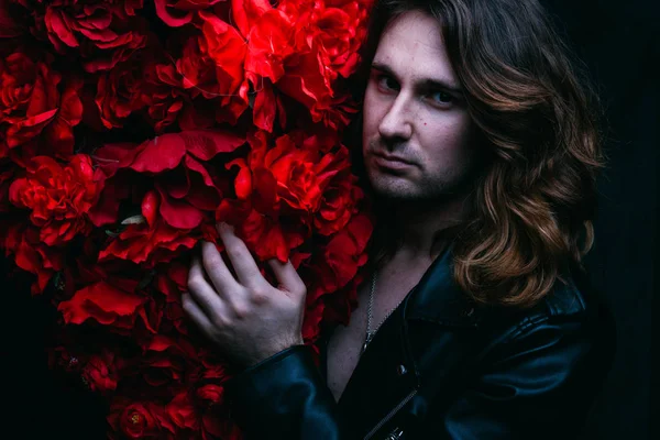 acımasız adam uzun saçlı çıplak bir vücut üzerinde deri ceketli kırmızı çiçek çiçekli dekor yakın duruyor. güç ve güzellik. Güzellik korumak. Saç Bakımı.